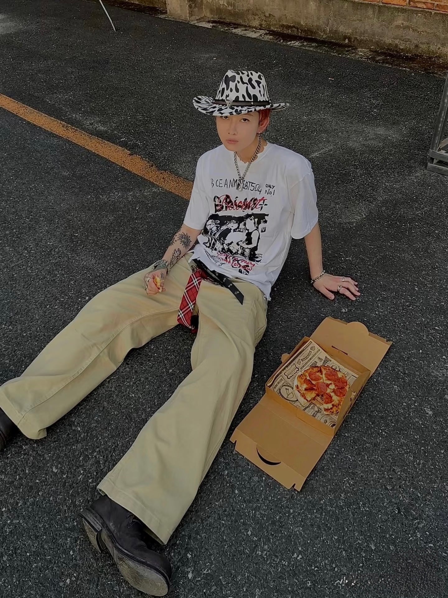 Person sitting on the ground with a pizza box next to them, wearing a hat and casual clothing.