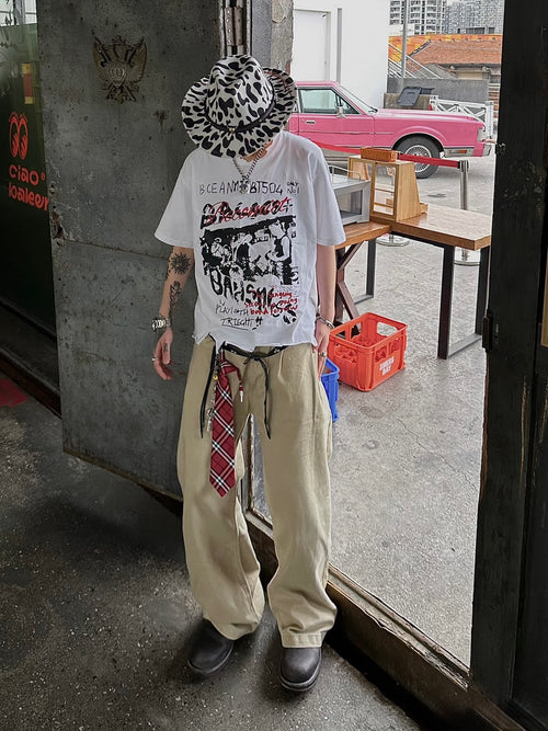 Person wearing a white graphic t-shirt, beige pants, and a patterned hat in an urban setting.