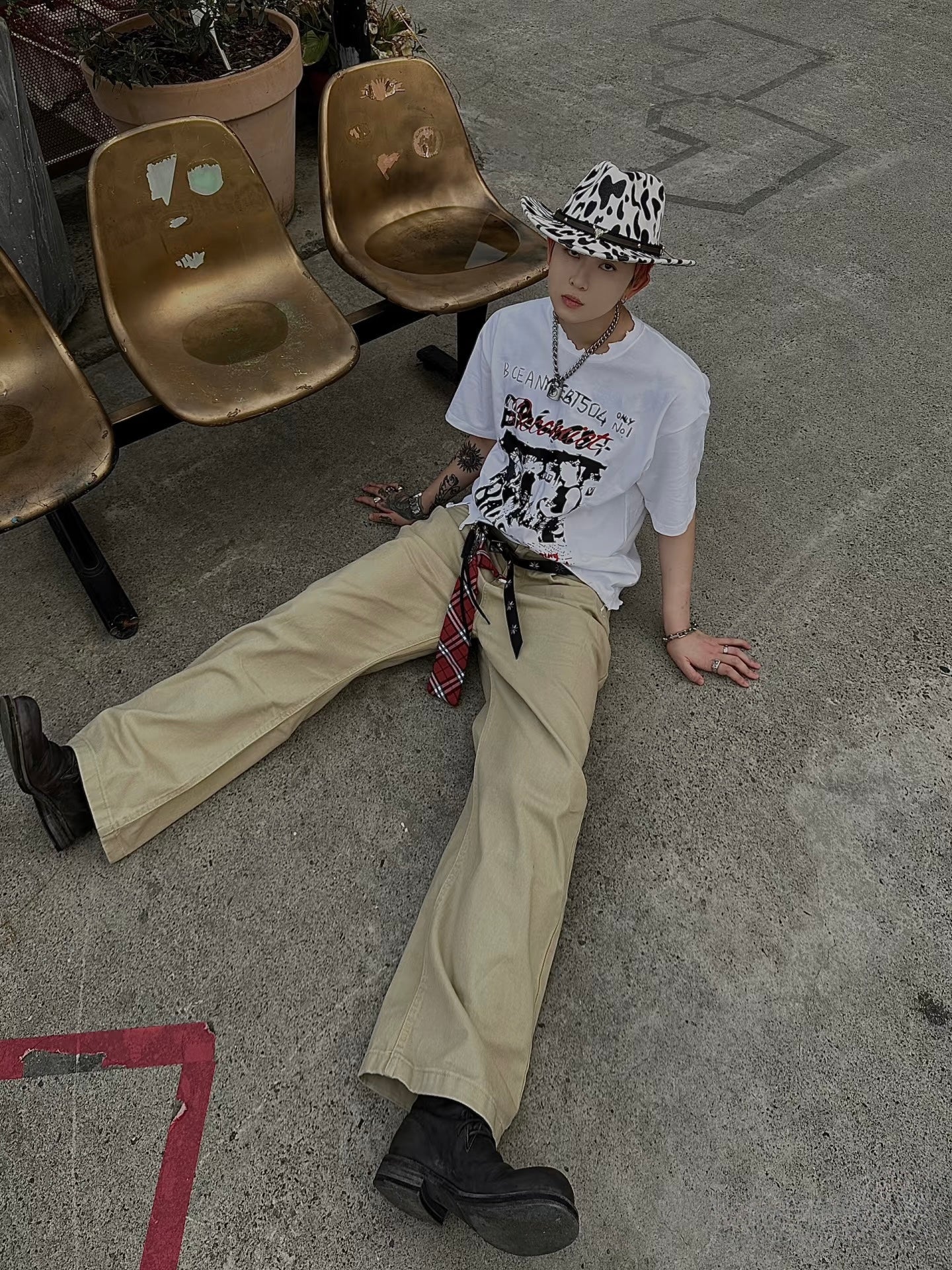 Person sitting on the ground wearing a white graphic t-shirt, beige pants, and a patterned hat.