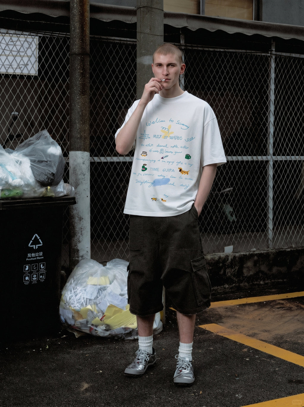 Man wearing a white graphic t-shirt and dark shorts standing in an outdoor setting with a fence and trash bins in the background.