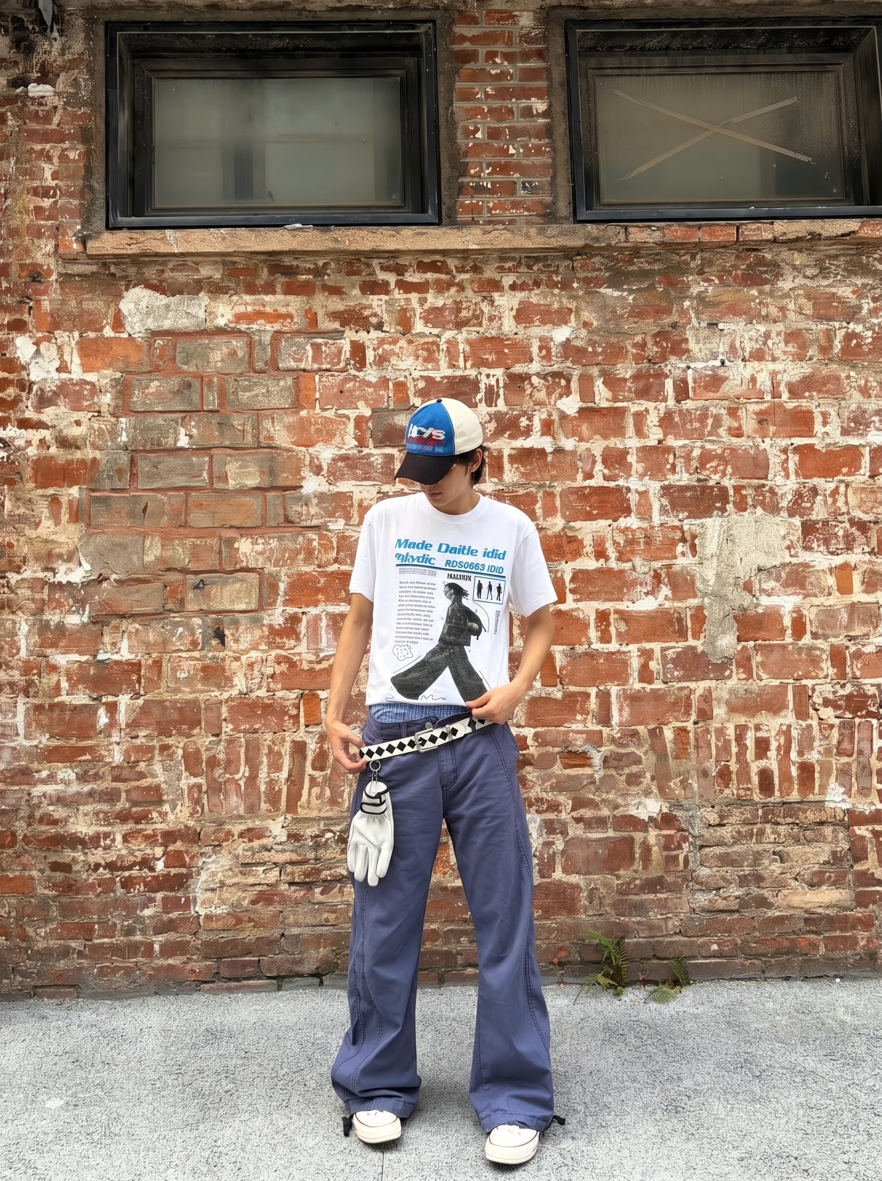 Person wearing a white t-shirt with black design, blue pants, and a cap standing against a brick wall.