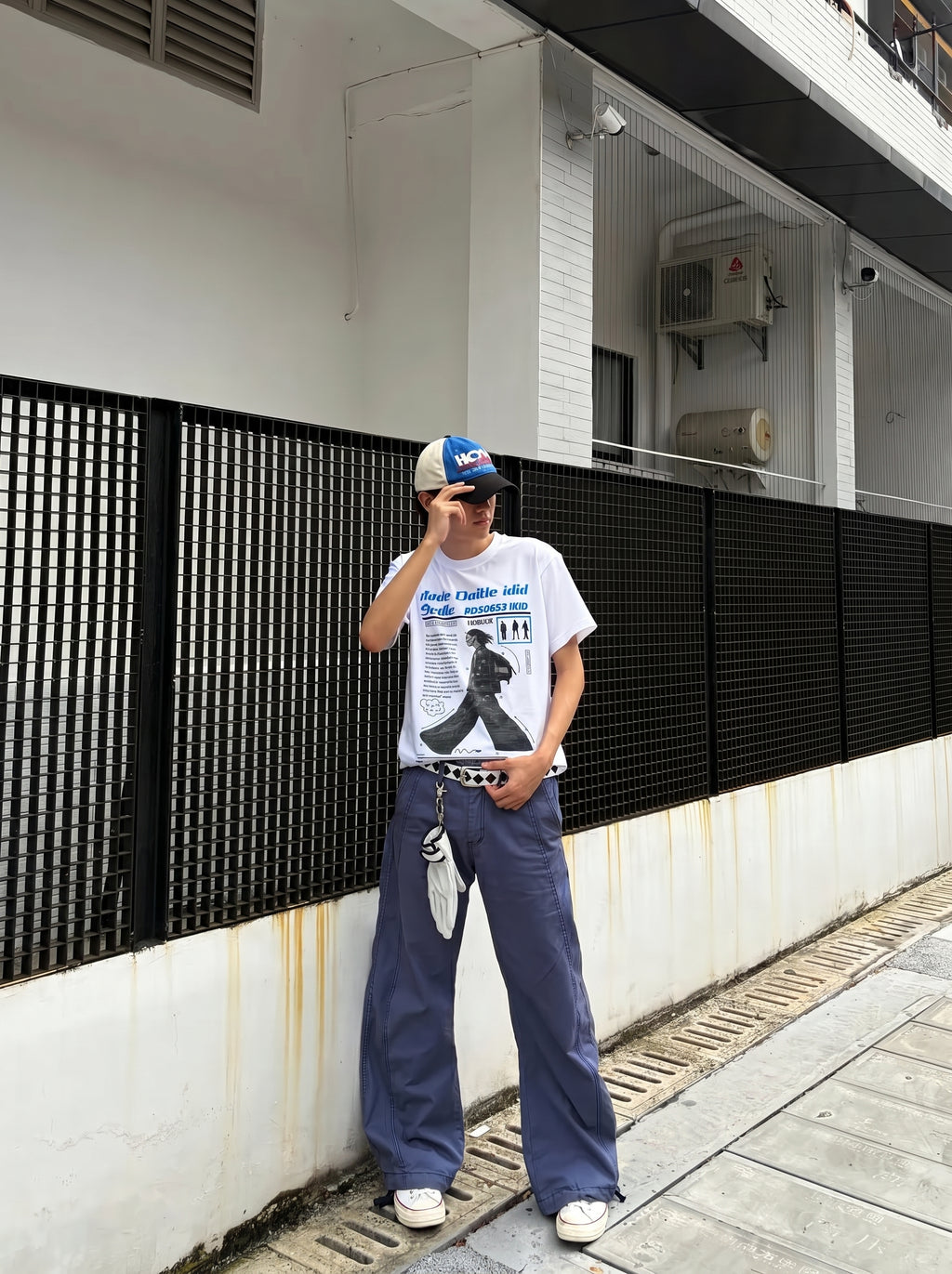 Person wearing a white t-shirt with a graphic design and blue jeans standing against a building wall.